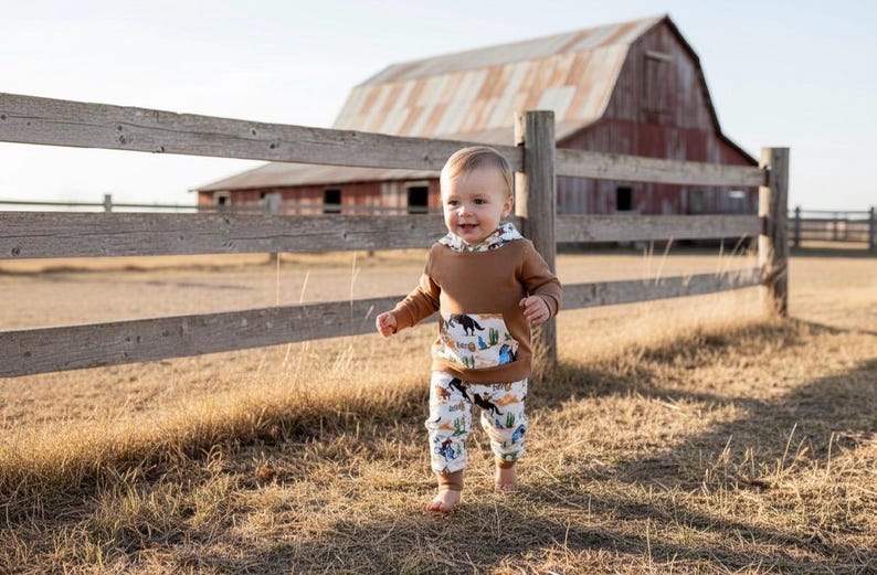 Puede incluir: Un ni&ntilde;o peque&ntilde;o con una sudadera con capucha marr&oacute;n con un bolsillo con una escena del desierto y pantalones a juego. El conjunto se encuentra frente a una valla de madera r&uacute;stica y un granero.