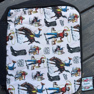 K&ouml;nnte beinhalten: Ein rechteckiges Etui mit schwarzem Rei&szlig;verschluss und einem wei&szlig;en Stoffeinsatz, bedruckt mit einem sich wiederholenden Muster aus Cowboys auf Pferden, lasso-werfenden Figuren, schwarzen Cowboystiefeln sowie dem Text "Yee Haw!" und "Giddy Up!".