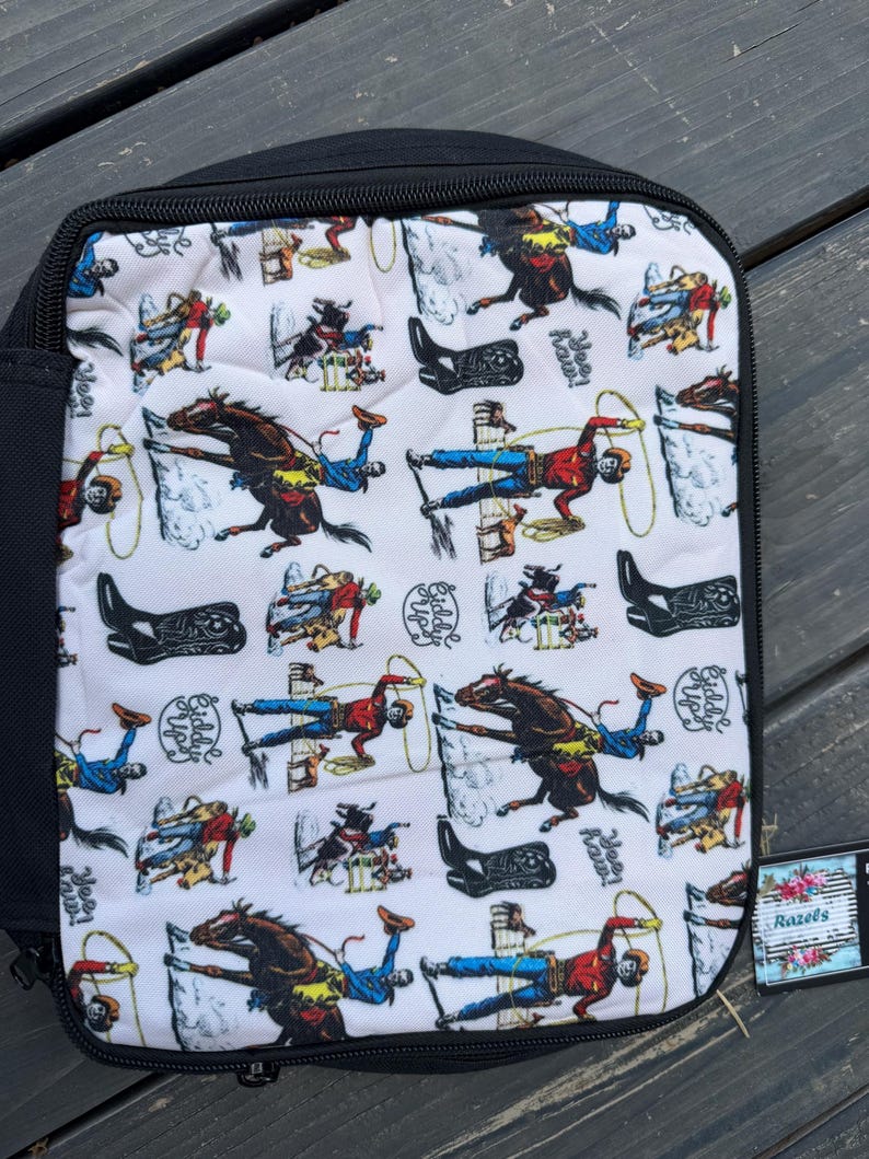 K&ouml;nnte beinhalten: Ein rechteckiges Etui mit schwarzem Rei&szlig;verschluss und einem wei&szlig;en Stoffeinsatz, bedruckt mit einem sich wiederholenden Muster aus Cowboys auf Pferden, lasso-werfenden Figuren, schwarzen Cowboystiefeln sowie dem Text "Yee Haw!" und "Giddy Up!".