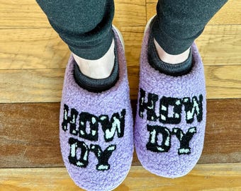 Könnte beinhalten: Ein Paar violette, flauschige Hausschuhe mit dem Wort "HOWDY" in Schwarz auf jedem Schuh bestickt.