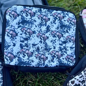 K&ouml;nnte beinhalten: Eine quadratische Lunchtasche mit einem sich wiederholenden Muster aus braunen bockenden Pferden und Reitern auf einem wei&szlig;-schwarzen Kuhfell-Hintergrund. Die Tasche hat einen schwarzen Rei&szlig;verschluss und einen schwarzen Stoffrand an den Kanten.