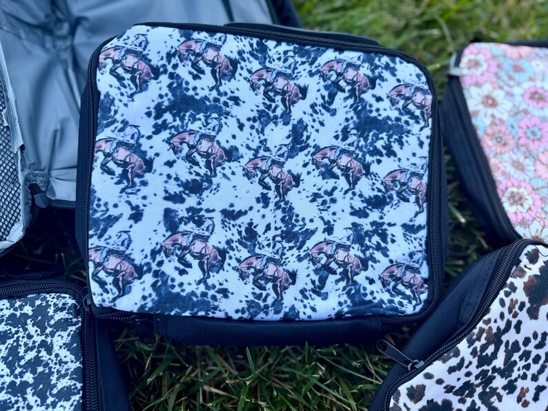 K&ouml;nnte beinhalten: Eine quadratische Lunchtasche mit einem sich wiederholenden Muster aus braunen bockenden Pferden und Reitern auf einem wei&szlig;-schwarzen Kuhfell-Hintergrund. Die Tasche hat einen schwarzen Rei&szlig;verschluss und einen schwarzen Stoffrand an den Kanten.