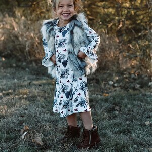 May include: A young girl wearing a white dress with a deer print and a gray faux fur vest. She is standing in a grassy area with trees in the background.
