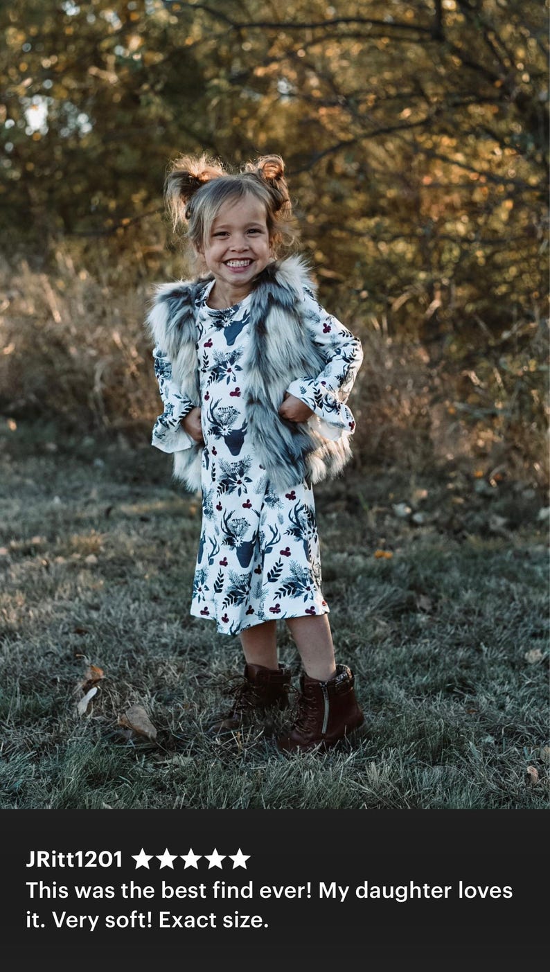May include: A young girl wearing a white dress with a deer print and a gray faux fur vest. She is standing in a grassy area with trees in the background.