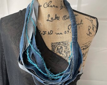 May include: A blue denim necklace made from strips of recycled denim fabric. The necklace is frayed and has a rustic look.