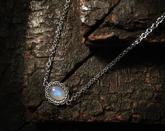 May include: A silver chain necklace featuring a central oval moonstone cabochon set in a decorative, scalloped metal frame. The gemstone displays a soft, iridescent blue glow against the polished silver setting and the delicate link chain.