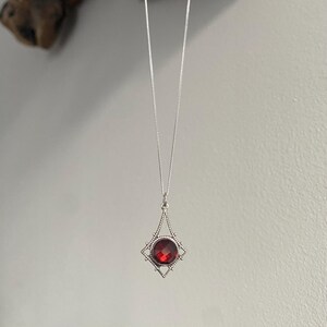 May include: A silver necklace with a pendant featuring a round, faceted, red gemstone. The pendant is set within a decorative, openwork frame. The necklace hangs against a neutral background.