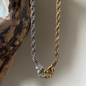 May include: A silver and gold chain necklace with two hands clasped together as the pendant. The hands are intertwined and facing each other.