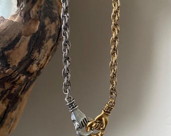 May include: A silver and gold chain necklace with two hands clasped together as the pendant. The hands are intertwined and facing each other.