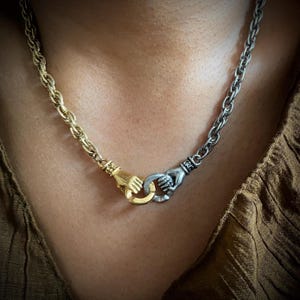May include: A silver and gold chain necklace with two clasped hands as the pendant.