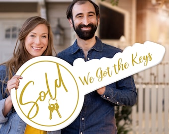 May include: A couple holds a large white key-shaped sign that says "Sold" in a gold circle and "We Got the Keys" on the handle. The sign is a real estate prop.