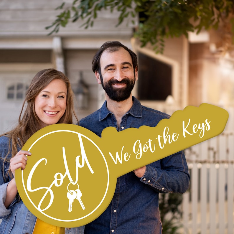 May include: A couple holds a large, gold key-shaped sign that says "Sold" on one side and "We got the keys" on the other. The sign is decorated with a pair of keys.