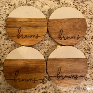 May include: Four round wooden coasters with a white top and a brown bottom. The word "browns" is engraved on each coaster.