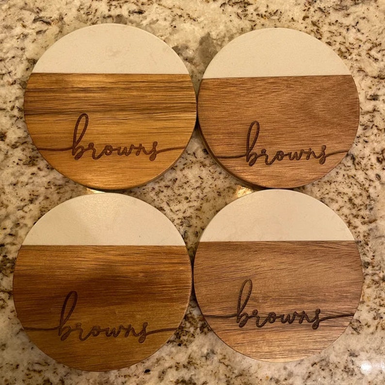 May include: Four round wooden coasters with a white top and a brown bottom. The word "browns" is engraved on each coaster.