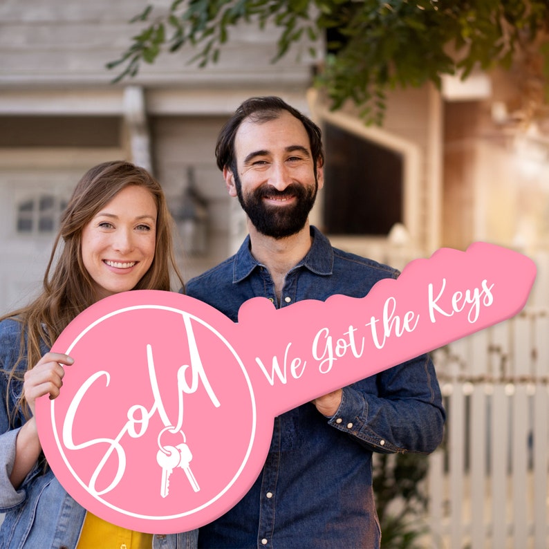 May include: A couple holds a large pink key-shaped sign that says "Sold" on one side and "We Got the Keys" on the other side. The sign is decorated with a white outline and white text.