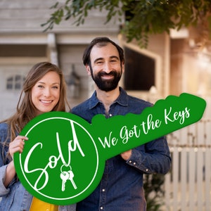 May include: A couple holds a large green key-shaped sign that says "Sold" on one side and "We got the keys" on the other side. The sign is decorated with white lettering and a pair of keys.