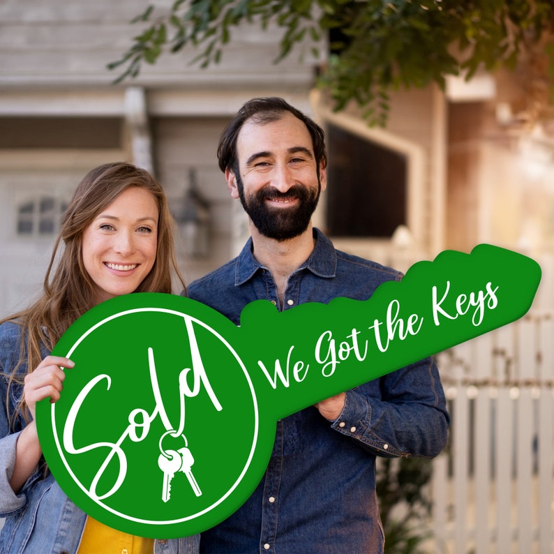 May include: A couple holds a large green key-shaped sign that says "Sold" on one side and "We got the keys" on the other side. The sign is decorated with white lettering and a pair of keys.