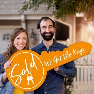 May include: A couple holds a large orange key-shaped sign that says "Sold" on one side and "We got the keys" on the other side. The sign is made of wood and has a white outline.