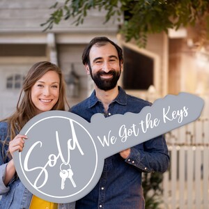 May include: A couple holds a large gray key-shaped sign. The sign says "Sold" on one side and "We got the keys" on the other side. The couple is smiling and standing in front of a house.