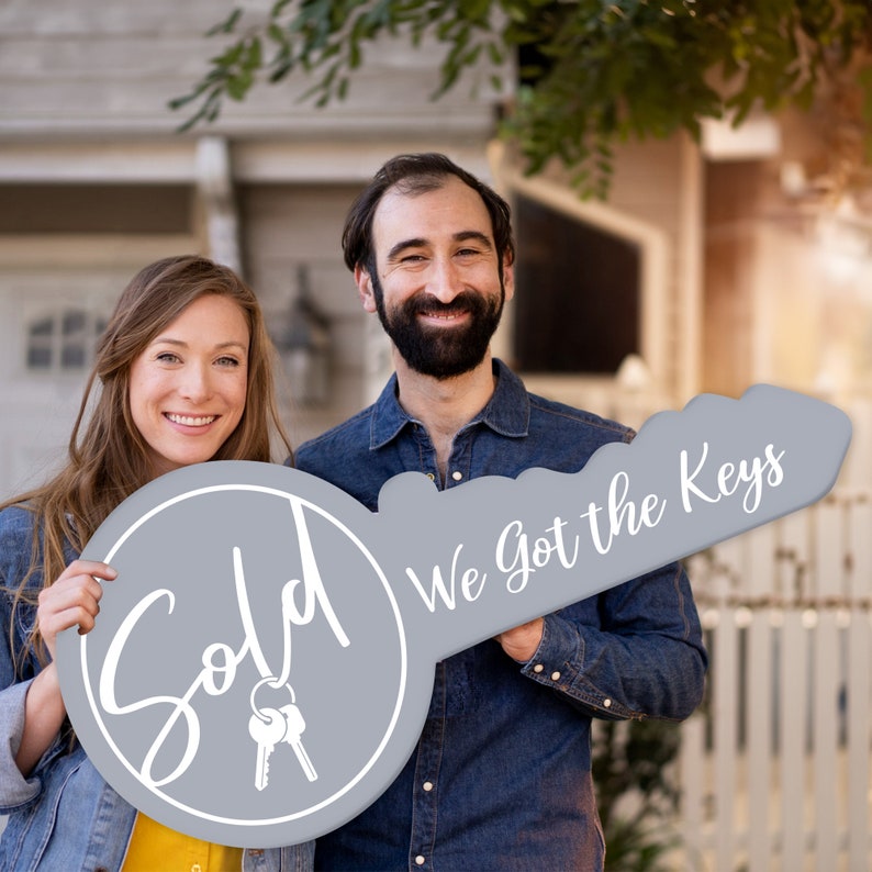 May include: A couple holds a large gray key-shaped sign. The sign says "Sold" on one side and "We got the keys" on the other side. The couple is smiling and standing in front of a house.