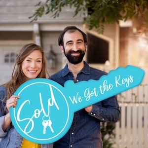 May include: A couple holds a large blue key-shaped sign that says "Sold" on one side and "We got the keys" on the other. The sign is held in front of a house.