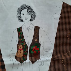 Op de afbeelding: Een wit stoffen paneel met een zwart-witte lijntekening van een persoon die een bruin-wit geruit vest draagt met geborduurde details. Het vest heeft een kat, een bij en een mand met bessen. Het stoffen paneel heeft de tekst "DreamSpinners" onderaan.