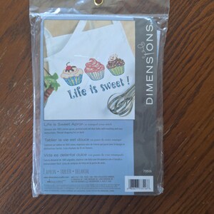 May include: A Dimensions brand stamped cross stitch kit for a white apron featuring three colorful cupcakes and the text Life is sweet! along with a whisk illustration. The kit is packaged in a clear plastic bag with blue and black branding.
