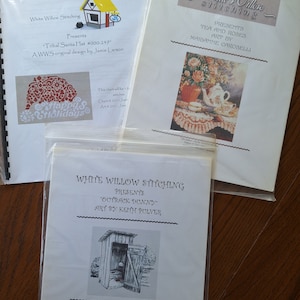 Könnte beinhalten: Drei weiße Papiermuster für Kreuzstich-Stickerei. Das erste Muster ist für eine Stammes-Santa-Mütze, das zweite für ein Tee- und Rosen-Design und das dritte für ein Outback-Klo. Jedes Muster enthält den Namen des Designs, den Namen des Künstlers und die Anzahl der Stiche.