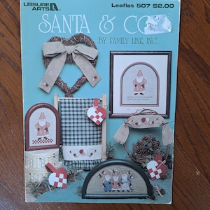 May include: A cross-stitch pattern booklet featuring Santa Claus and other Christmas characters. The booklet is titled "Santa & Co. by Family Line, Inc." and includes a variety of designs, including a wreath, a towel, and a stocking.