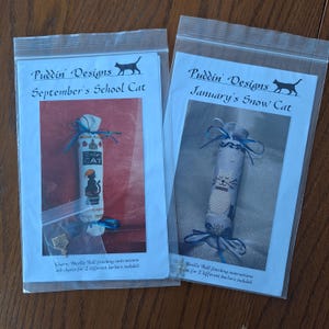 May include: Two clear plastic bags containing cross-stitch kits. The left bag features a design labeled "September's School Cat" with a black cat illustration. The right bag is labeled "January's Snow Cat". Both kits include instructions and charts.