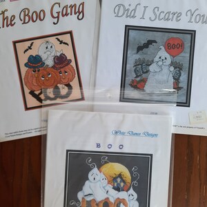 May include: Three cross stitch patterns featuring Halloween themes. The first pattern is titled "The Boo Gang" and features a group of ghosts and pumpkins. The second pattern is titled "Did I Scare You?" and features a ghost holding a red balloon with the word "Boo!" on it. The third pattern is titled "Boo" and features three ghosts in front of a moon with a house behind them.