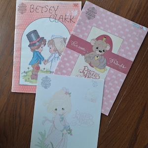 Puede incluir: Tres patrones de punto de cruz con personajes de Precious Moments. El primer patrón muestra una pareja con atuendo de boda, el segundo muestra un oso sosteniendo un corazón y el tercero muestra una niña con un vestido blanco sosteniendo flores. Todos los patrones están sobre fondos rosa y blanco.