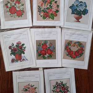 Puede incluir: Una colección de ocho gráficos de punto de cruz que presentan diseños florales. Los gráficos están impresos en papel blanco con un fondo rosa claro y presentan varias flores, incluidas rosas, lirios e hortensias.