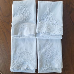Puede incluir: Cuatro lienzos rectangulares blancos dispuestos en cuadrícula, cada uno con un bordado blanco sobre blanco distinto. Los diseños incluyen patrones florales complejos, detalles de encaje calado y bordes festoneados en las telas.