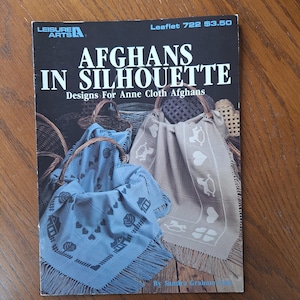 Op de afbeelding: Een boekomslag getiteld "AFGHANS IN SILHOUETTE" met ontwerpen voor Anne Cloth Afghans. De omslag toont afbeeldingen van afgewerkte afghanen in manden, met een blauwe en een bruine afghaanse deken. Het boek is van Leisure Arts.