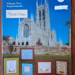 Op de afbeelding: Een boek getiteld "Our Best to You, Volume Two Inspirationals" door Needles 'n Hoops. De omslag toont een foto van een grote stenen kerk met een blauwe lucht en witte wolken. Het boek bevat vier kruissteekpatronen met inspirerende citaten.