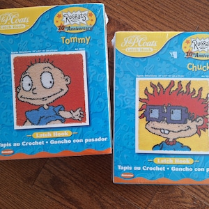 May include: Two Rugrats latch hook kits featuring Tommy and Chuckie. The kits are blue and orange with the Rugrats logo and 10th Anniversary text. The kits are approximately 12 inches by 16 inches.