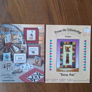 May include: Two cross stitch pattern books with Native American themes. The first book is titled "Tribal Designs from Weavings & Beadwork" and features a variety of designs, including a Native American woman, a beaded bag, and a rug. The second book is titled "Cross the Lilliebridge Designs" and features a totem pole design.