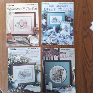 May include: Four Leisure Arts cross-stitch pattern leaflets by Paula Vaughan titled Reflections Of The Past, Sweet Violets, Poems and Promises, and The Quilter. Each booklet features a color photograph of a finished needlework project.