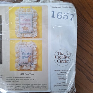 May include: A cross-stitch kit for two baby-themed wall hangings. Each features a stitched design with the phrases "Baby's Napping" and "I'm Awake Now". The finished size is approximately 13.3 cm x 17.7 cm with a lace trim.