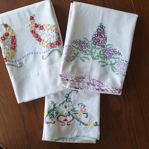 May include: Three white pillowcases with colorful floral embroidery. One pillowcase features red, yellow, and green flowers. Another has purple flowers with green leaves and a lace trim. The third has a floral design with pink and yellow flowers.