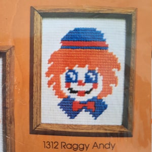 May include: A framed cross-stitch pattern of a Raggy Andy clown with orange hair, a blue hat, and a red bow tie. The pattern is labeled "1312 Raggy Andy".