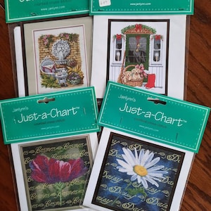 May include: Four different counted cross stitch patterns from Janlynn's Just-a-Chart collection. The patterns include a floral design with a red flower, a daisy with a yellow centre, a welcome sign with a basket of puppies, and a fountain with a stone statue.