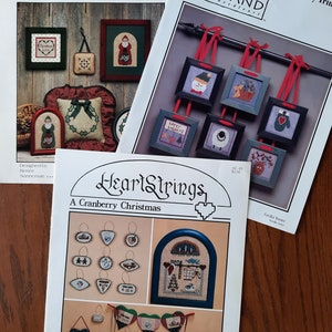 May include: Three different cross-stitch patterns for holiday ornaments. The first pattern is from The Need'l Love Company and features a Santa Claus and a wreath. The second pattern is from Heart in Hand Needleart and features a snowman, a sheep, a bird, and a mitten. The third pattern is from HeartStrings and features a variety of Christmas ornaments, including a rocking horse, a Santa Claus, and a heart.