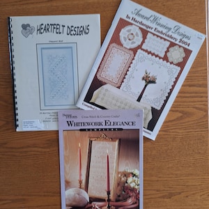 May include: Three instructional craft booklets featuring embroidery and cross-stitch patterns. The booklets are titled Heartfelt Designs, Award-Winning Designs In Hardanger Embroidery 2004, and Whitework Elegance, each displaying various needlework projects.