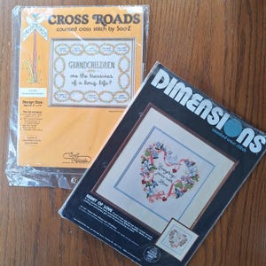 May include: Two cross stitch patterns, one from Cross Roads and one from Dimensions. The Cross Roads pattern is in a plastic bag and features a wreath with the text "Grandchildren are the treasures of a long life!" The Dimensions pattern is in a black frame and features a heart with flowers and the text "Everything grows with love".