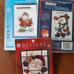Puede incluir: Tres kits de punto de cruz contados con temas de invierno y Navidad. Un kit muestra un muñeco de nieve con el texto "Snow Much Fun". Otro kit tiene un muñeco de nieve con el texto "Being Perfect". El tercer kit presenta a Papá Noel y un reno.