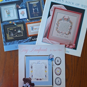 Puede incluir: Tres libros de patrones de punto de cruz con cubiertas azules, rosas y blancas. Los títulos son "Unique Birth Announcements", "A Baby's Lock of Hair" y "Tiny Handprints".