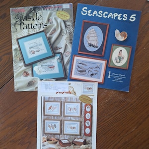 May include: Three softcover craft pattern booklets featuring nautical themes. The top left booklet is titled Seaside Patterns. The top right booklet is titled SEASCAPES 5. The bottom booklet is titled sea shells. Each cover displays cross-stitch designs.