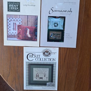 May include: Three cross-stitch pattern booklets. The top left booklet is titled "Long Stockings" by Bent Creek. The top right booklet is by Samsarah Design Studio. The bottom booklet is "The Ricket Collection" with a folk nativity scene.